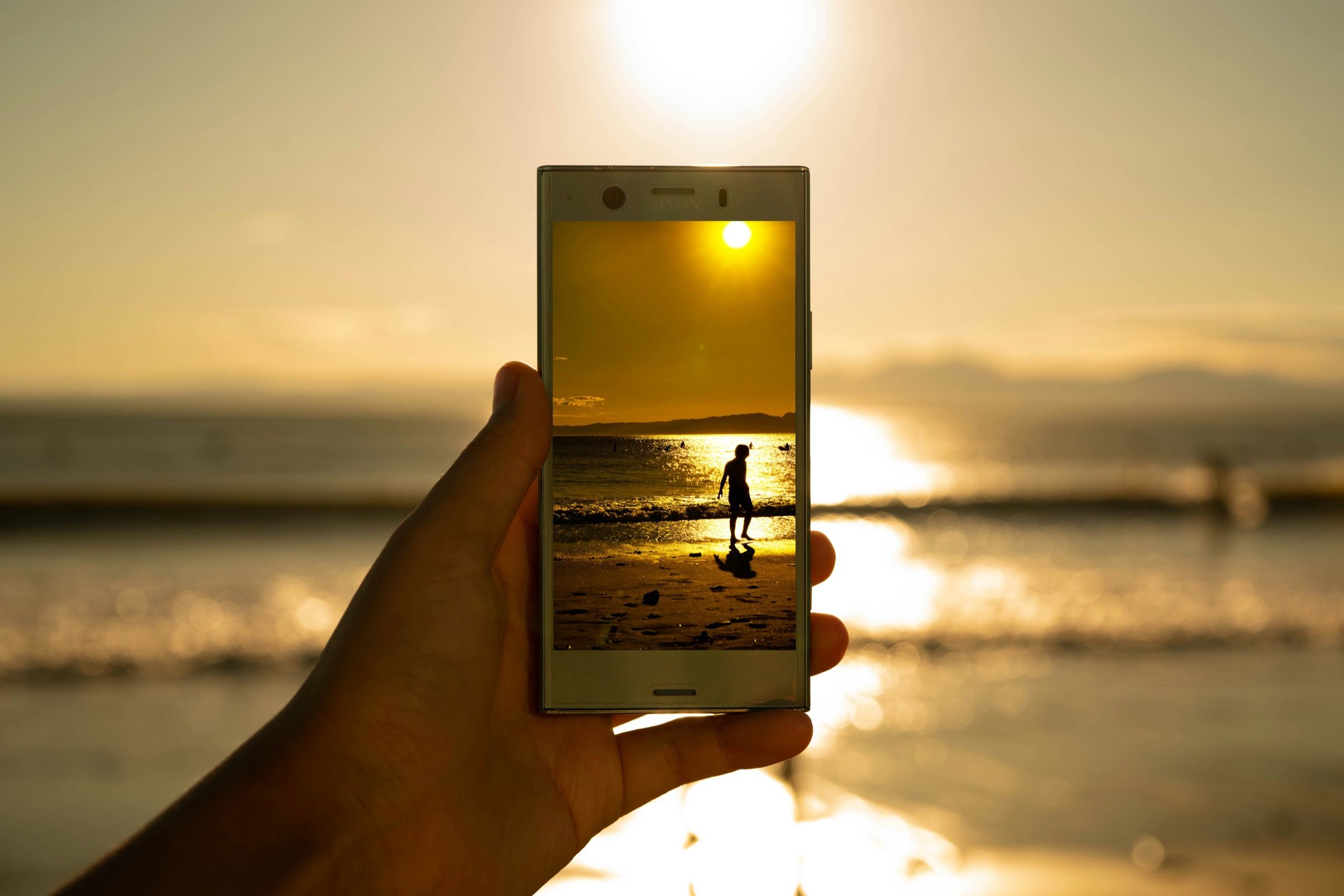 A hand holds up a phone against a golden sunset over the sea, its screen showing the silhouette of a person on the beach.