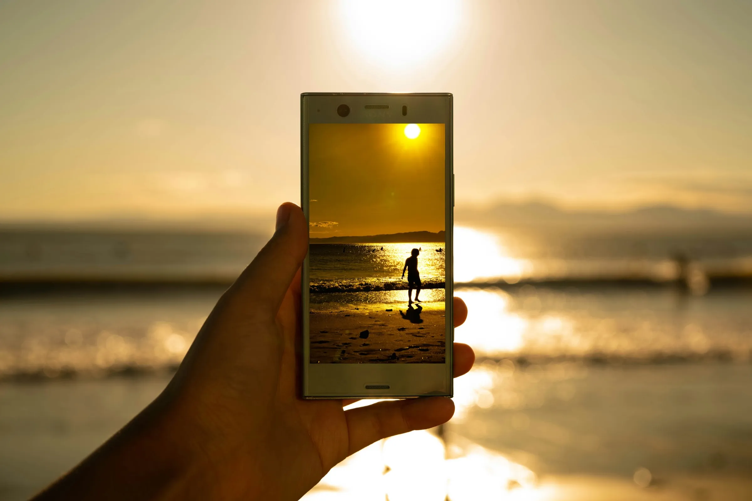A hand holds up a phone against a golden sunset over the sea, its screen showing the silhouette of a person on the beach.