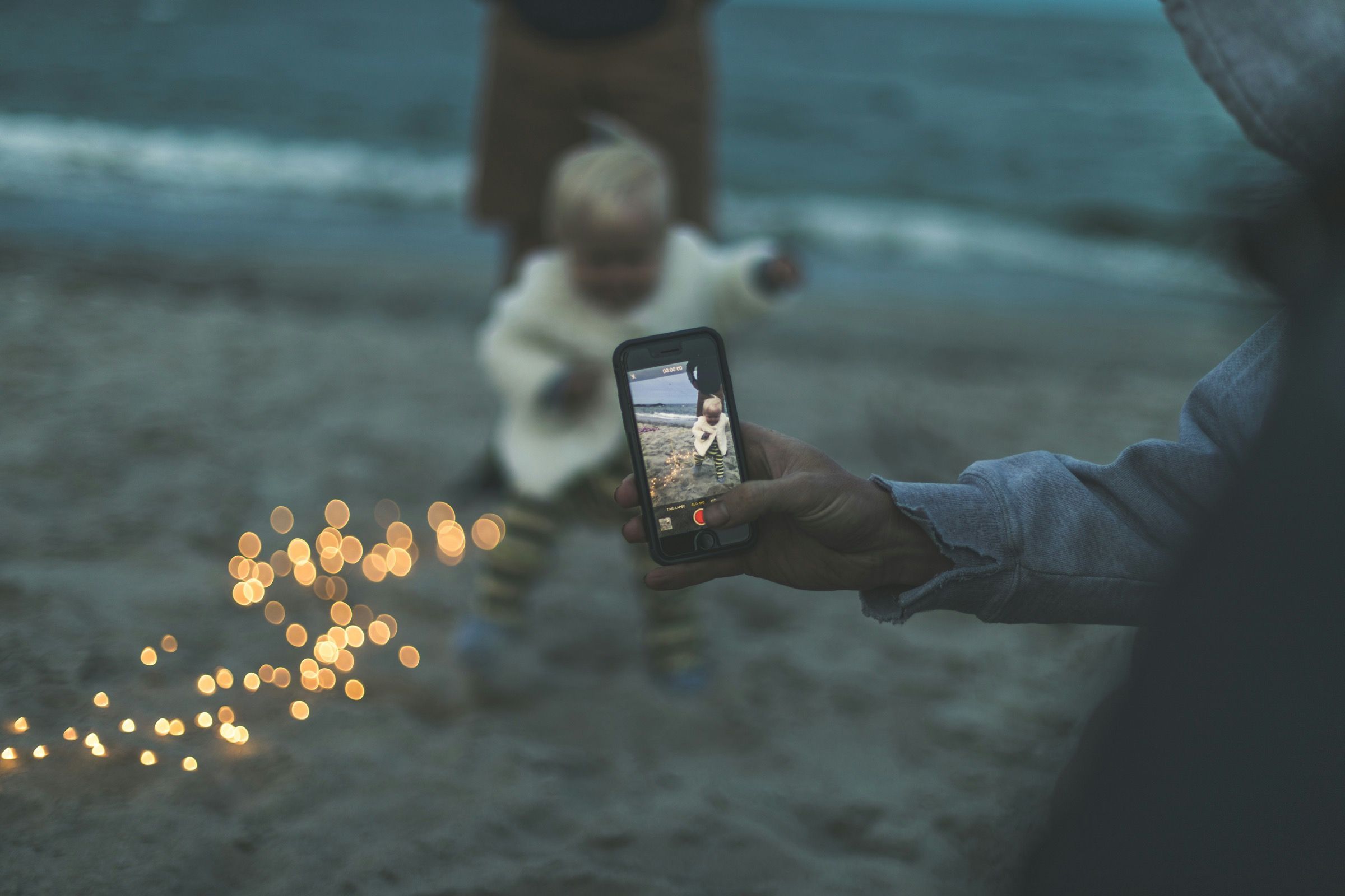 Close na mão de um pai filmando o filho pequeno no celular numa praia ao entardecer, com o rosto da criança fora de foco e luzes douradas ao fundo.
