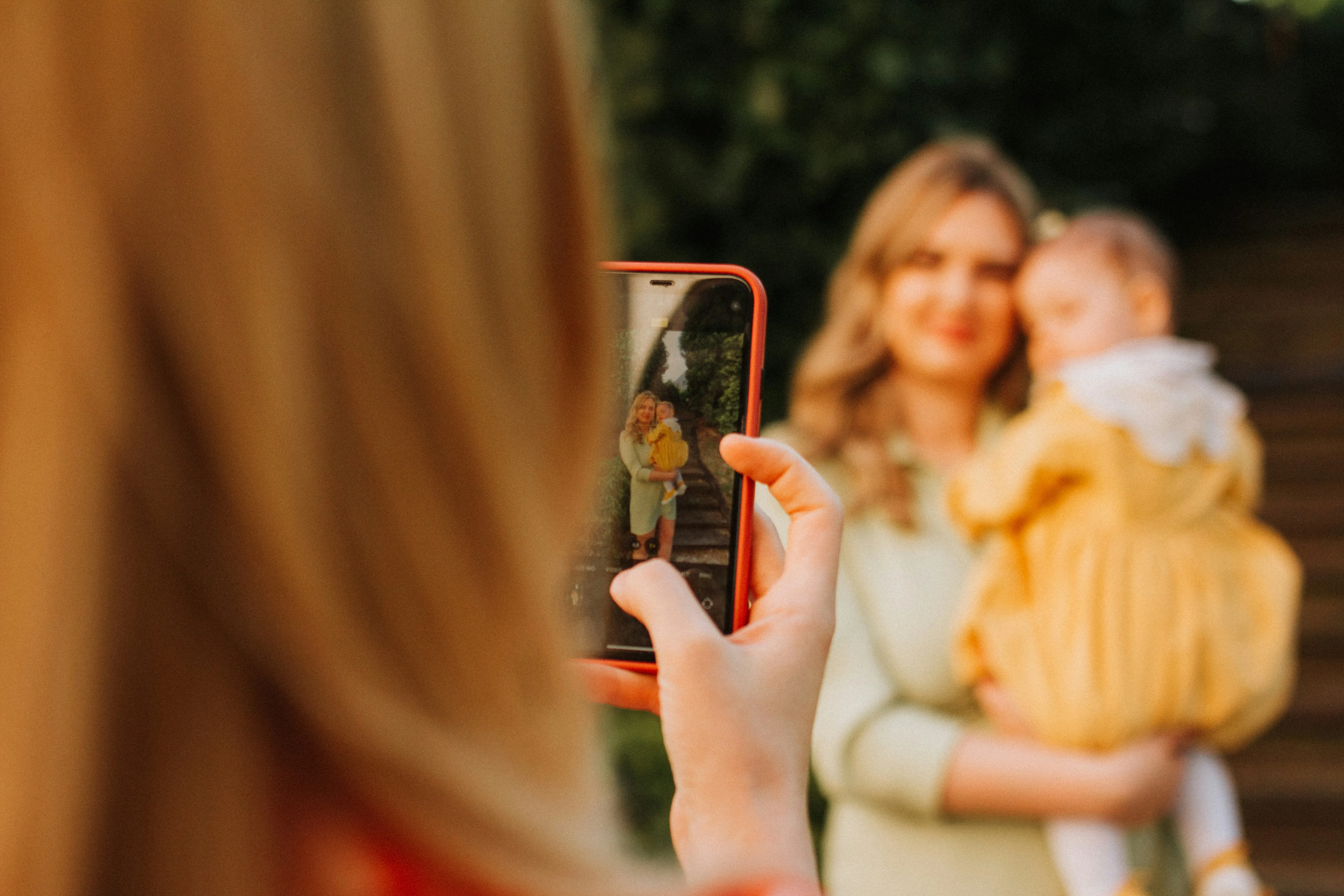 Seen over a shoulder, a hand holds a phone photographing a mother holding her baby in warm evening light.